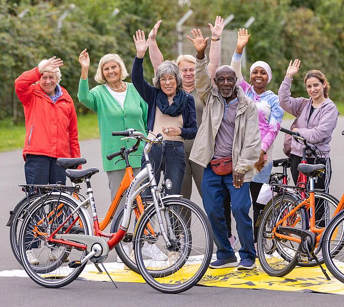 Eine Gruppe steht auf einer asphaltierten Fläche und winkt freundlich in die Kamera. Vor ihnen stehen drei orangefarbene Fahrräder. Die Gruppe wirkt fröhlich und gemeinschaftlich, im Hintergrund ist Grünbewuchs und ein Zaun zu sehen.﻿