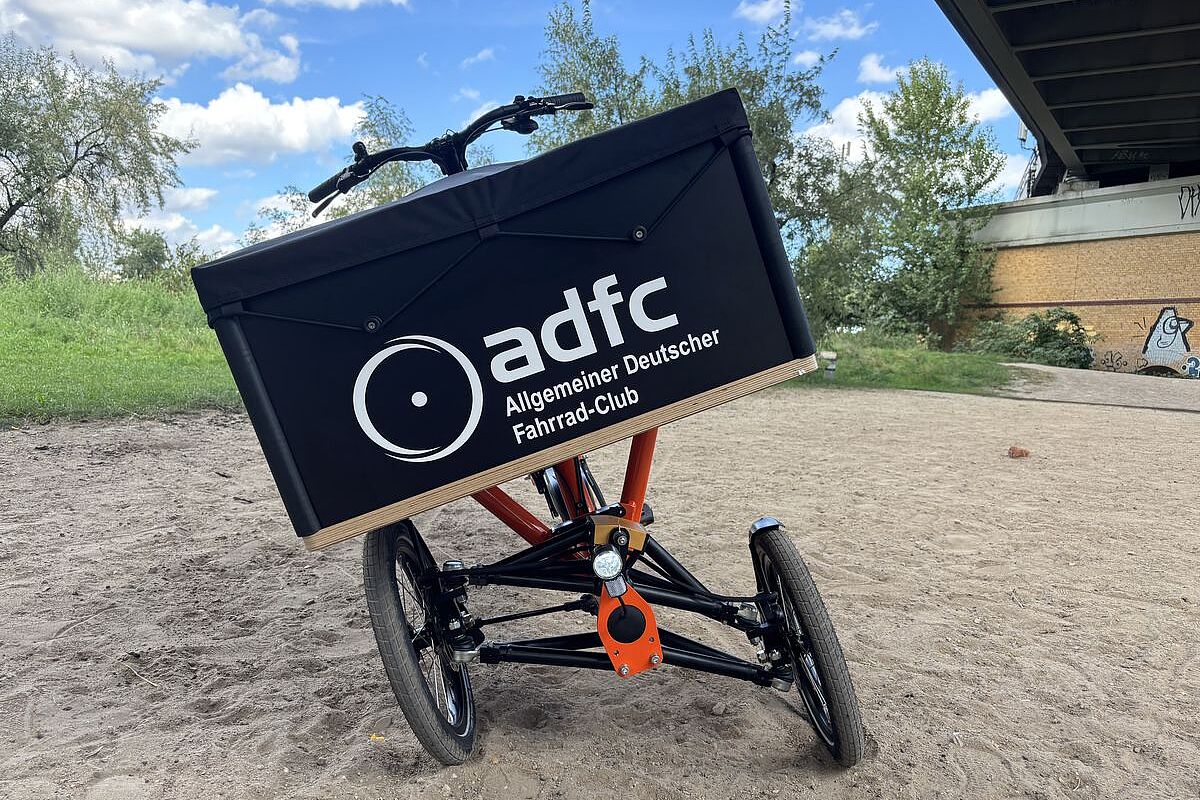Ein dreirädriges Lastenrad mit schräg geneigter Transportbox steht auf einem sandigen Weg.